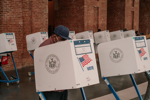 A Black man votes at a privacy booth at Brooklyn Museum's voting site; a brick background can be seen